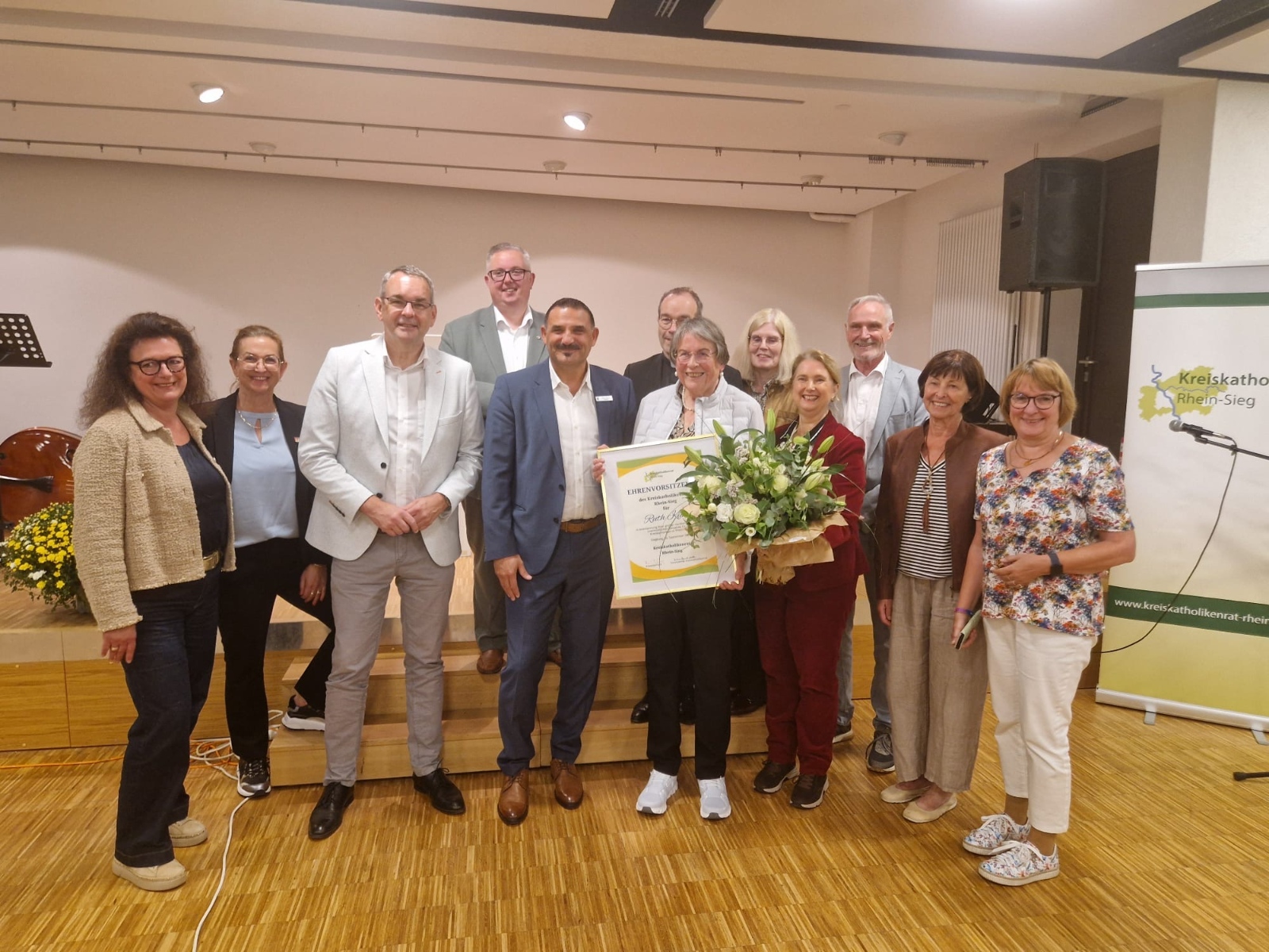 Das Foto – Quelle: Dr. Ralf Forsbach – zeigt die Ehrenvorsitzende des Kreiskatholikenrats Rhein-Sieg, Ruth Kühn (Mitte mit Urkunde) gemeinsam mit Kreisdechant Hans-Josef Lahr (dahinter), Bettina Heinrichs-Müller, Vorsitzende Kreiskatholikenrat (1.R., 3.v.r.), stellv. Landrat Erkan Zorlu (1.R., 4.v.l.), Bürgermeister Stefan Rosemann (1.R.,3.v.r.), Barbara Lülsdorf, stellv. Bürgermeisterin Niederkassel (1.R., 2.v.l.), Claudia Feld-Wielpütz, stellv. Bürgermeisterin Sankt Augustin (1.R., 1.v.l.), Sascha Lienesch MdL (2.R., 1.v.l.), Prof. Dr. Ursula Nothelle-Wildfeuer (2.R., 2.v.r.), Dr. Oliver Funken, stellv. Vorsitzender Kreiskatholikenrat (2.R., 1.v.r.), Elisabeth Bungartz, Vorstandsmitglied (1.R., 2.v.r.), Elisabeth Becker, Vorstandsmitglied (1.R., 1.v.r.).